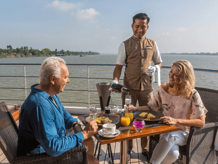 A man and woman being served a beverage by a butler on the outdoor terrace of the Scenic Spirit ship