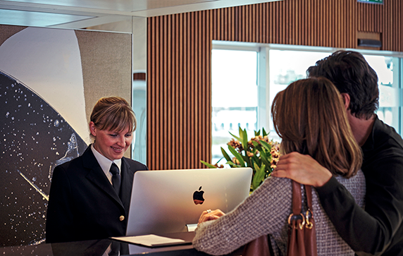  A couple at a reception desk being assisted by a smiling woman looking at a computer screen.  