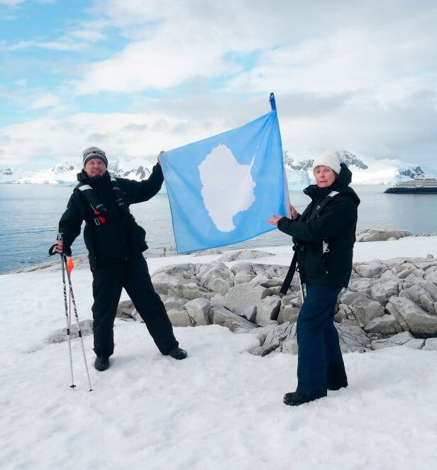 Two guests holding the Antarctica flag on a snowy landing with Scenic Eclipse in the background